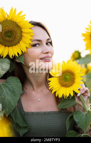 Modeporträt niedliche Frau schaut mit einem Lächeln in die Kamera und fröhlicher Ausdrucksform steht vor einem blühenden Sonnenblumenfeld Stockfoto