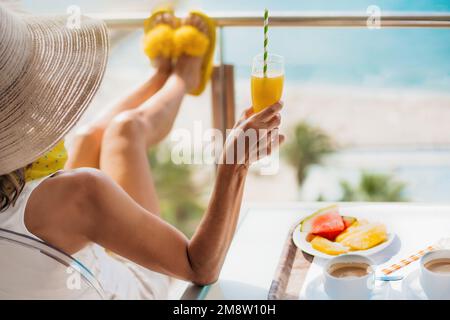 Detail einer Frau mittleren Alters mit Strohhut, die auf der Hotelterrasse frühstückt und das Meer betrachtet. Konzept von Entspannung und Sommer Stockfoto