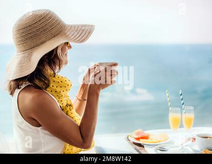 Eine Frau mit Strohhut hält eine Tasse Kaffee mit beiden Händen, während sie auf der Hotelterrasse auf das Meer blickt. Frühstückskonzept am Meer Stockfoto