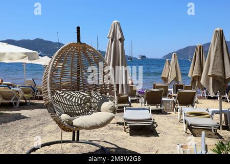 Korbstühle am Strand mit Sonnenschirmen und Liegestühlen. Sonniges Resort, Blick auf Berge und Yachten in der Ferne Stockfoto