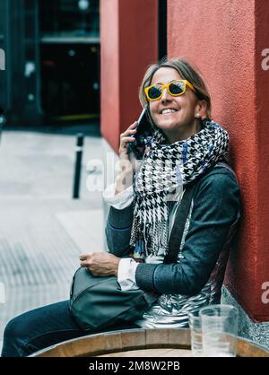 Eine Frau mittleren Alters, die ein Kopftuch und eine gelbe Sonnenbrille trägt, spricht am Telefon, während sie lächelt, vor einem roten Wandhintergrund. Stockfoto