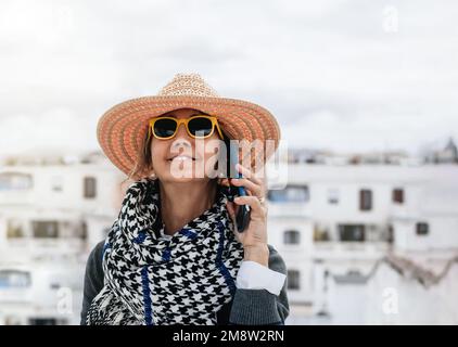 Vorderansicht eine Frau mittleren Alters, die einen Strohhut und eine Sonnenbrille trägt, lächelt, während sie auf einer Terrasse telefoniert. Stockfoto
