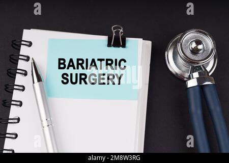 Medizinisches Konzept. Auf einer schwarzen Oberfläche, einem Stethoskop, einem Notizbuch, einem Stift und einem blauen Aufkleber mit der Aufschrift - BARIATRISCHE CHIRURGIE Stockfoto