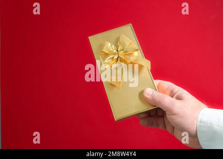 Ein goldenes Geschenk in der Hand eines Mannes mit Schleife. Valentinstag. Nahaufnahme auf rotem Hintergrund. Platz für Text Stockfoto