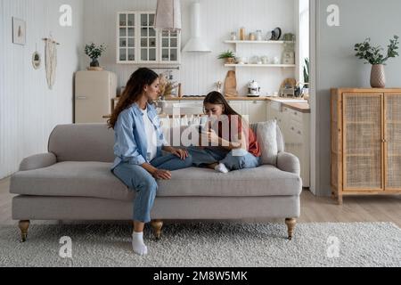 Trauriges Teenager-Mädchen, das mit der Mutter auf dem Sofa sitzt und ihr Handy in der Hand hält und in sozialen Medien gemobbt wird Stockfoto