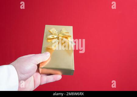 Ein wunderschönes Geschenk in einem goldenen Paket in der Hand eines Mannes mit Schleife. Nahaufnahme auf rotem Hintergrund. Platz für Text Stockfoto