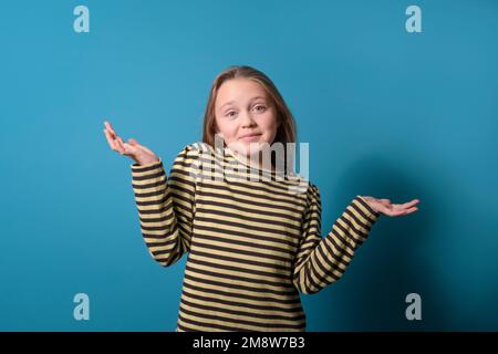 Ich weiß es nicht. Teenager-Mädchen. Studioporträt auf blauem Hintergrund. Gelb gestreiftes Hemd mit langen Ärmeln. Lange Haare. Sarkasmus Stockfoto