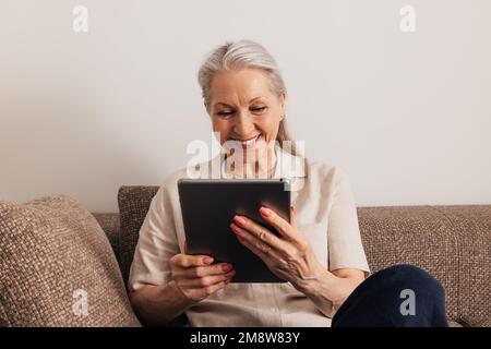 Nahaufnahme einer lächelnden Seniorin mit grauem Haar, die ein digitales Tablet hält, während sie auf dem Sofa sitzt Stockfoto