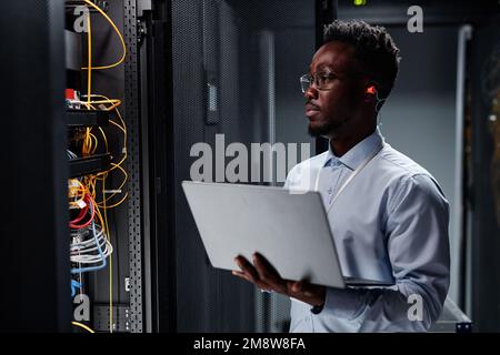 Porträt eines Schwarzen als Netzwerktechniker im Serverraum mit einem Laptop, minimal Stockfoto