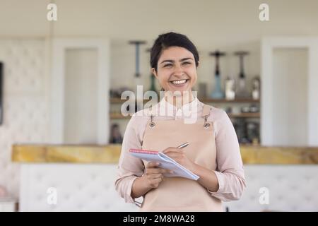 Glückliche, wunderschöne indische Kellnerin in Uniform Schürze, die einen Stift hält Stockfoto