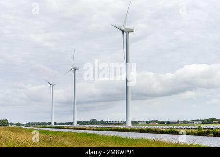 Windturbinen entlang eines Kanals in der Landschaft der Niederlande an einem bewölkten Sommertag. Reihen von Solarpaneelen befinden sich am Fuße der Turbinen. Stockfoto