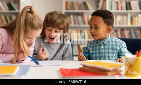 Zeit für die Schule. Multiethnische Kinder, die in Notizbüchern schreiben und lächeln, miteinander reden und lachen, am Tisch sitzen Stockfoto