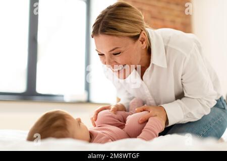 Kinderbetreuungskonzept. Glückliche Mutter macht Gymnastik für ihr Neugeborenes, verbringt Zeit zusammen im Schlafzimmer Stockfoto