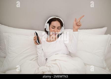 Fröhliche Brünette, die im Bett Musik hört, telefoniert Stockfoto