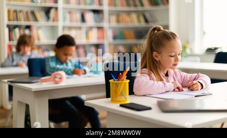 Verschiedene Kinder sitzen in der Grundschule oder im Kindergarten an Tischen und schreiben oder zeichnen in Notizbüchern Stockfoto