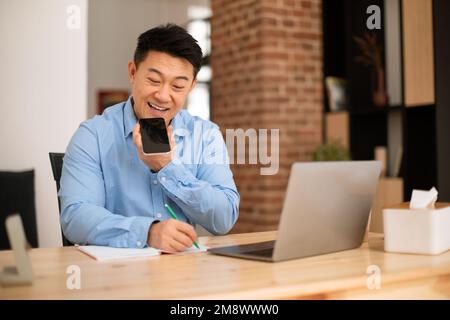 Anwendung für digitale Lautsprecher. Positiver asiatischer, reifer Geschäftsmann, der Sprachassistent am Handy benutzt, an einem Laptop arbeitet Stockfoto