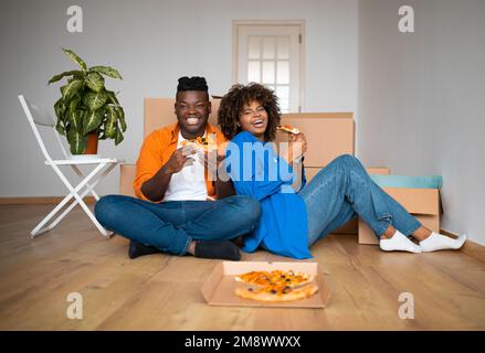 Schwarzes Paar, das auf dem Boden sitzt, Pizza isst, sich ausruht, nachdem er nach Hause gezogen ist Stockfoto