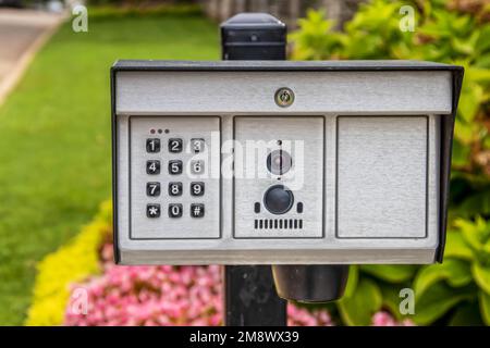 Elektrischer Toröffner mit Tastenfeld und Kamera und Mikrofon im Außenbereich des Anwesens mit wunderschönen verschwommenen Blumen im Hintergrund Stockfoto