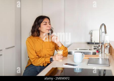 Junge Frau Sitzt An Der Küchentheke Und Isst Pudding Aus Plastikbecher Stockfoto