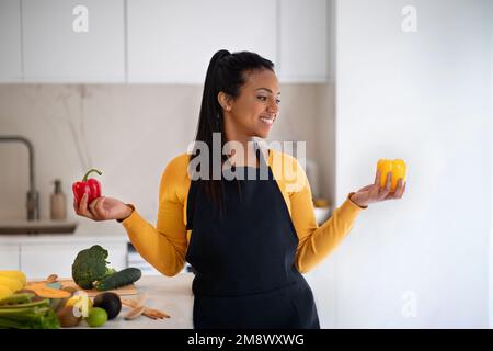Eine glückliche junge afroamerikanische Frau mit Schürze hält am Tisch roten und gelben Pfeffer mit Bio-Gemüse in den Händen Stockfoto