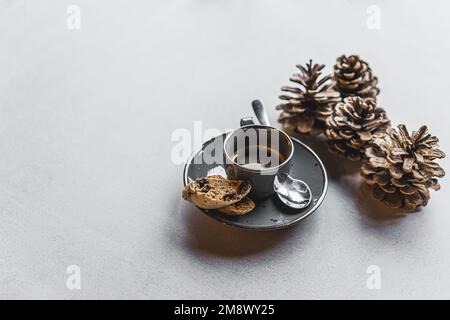 Kleine graue Tasse Espresso mit zwei Keksen neben dekorativen Kiefernzapfen auf einem Steintisch. Herbstsonnengetränk. Horizontalschuss. Hochwertiges Foto Stockfoto