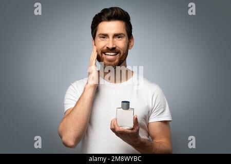 Grooming- und People-Konzept. Ein glücklicher, gutaussehender, weißer Mann, der Parfüm auf dem Bart aufträgt, grauer Studiohintergrund Stockfoto