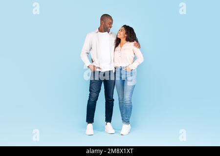 Lange Aufnahme eines schwarzen Paares, das sich umarmt und anschaut, auf blauem Hintergrund posiert, ganze Körperlänge Stockfoto