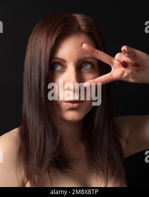 Studioporträt einer 40-jährigen Frau, die Sieg- oder Friedensgeste zeigt, mit den Fingern nahe dem Auge. Brünett mit langen Haaren und ausgestreckten Lippen, die wegschauen Stockfoto