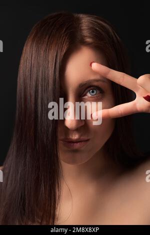 Porträt einer netten erwachsenen Dame, die ein Siegesschild nahe dem Auge zeigt und in die Kamera schaut. Reizende, brünette Frau mit langen Haaren, die ein Auge bedeckt Stockfoto