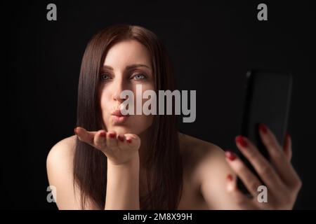 Eine Frau, die Lippen bläst, Luftküsse über die Handfläche schickt und Selfie in die Handykamera nimmt. 40s Frau mit langen Haaren, die flirtet und in die Kamera schaut Stockfoto