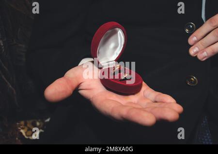 Zwei goldene Eheringe liegen in einer roten Schachtel auf der männlichen Hand des Bräutigams Stockfoto