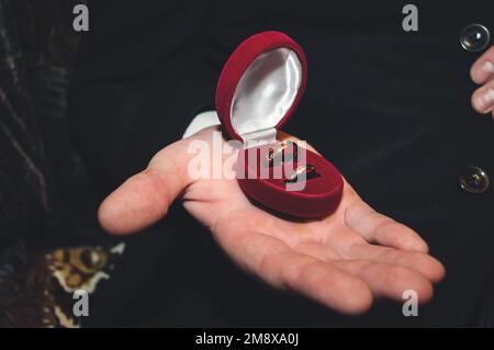 Zwei goldene Eheringe liegen in einer roten Schachtel auf der männlichen Hand des Bräutigams Stockfoto