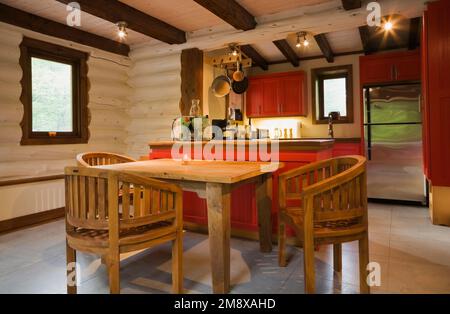 Hölzerner Frühstückstisch mit runden Lehnstühlen und Blick auf die Küche mit roten Holzschränken im 1. Stock im Blockhaus im skandinavischen Stil. Stockfoto