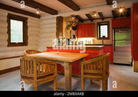 Hölzerner Frühstückstisch mit runden Lehnstühlen und Blick auf die Küche mit roten Holzschränken im 1. Stock im Blockhaus im skandinavischen Stil. Stockfoto