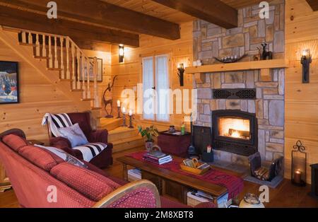 Burgunderrot mit weißem Polka-Dot-Polstersofa, Sessel mit Überwurf und Kissen, hölzerner Couchtisch und beleuchtetem Holzkamin aus Naturstein Stockfoto