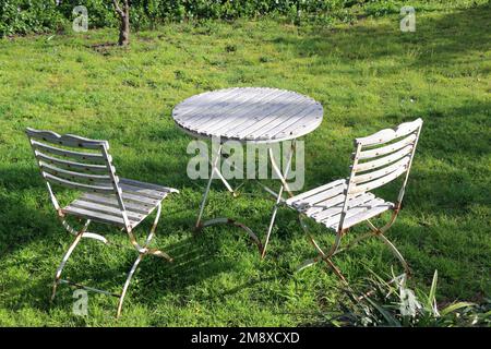 Verwitterte weiße Tische und Stühle in einem tiefgrünen Rasen bieten einen einladenden Ort zum Entspannen im Frühling. Stockfoto