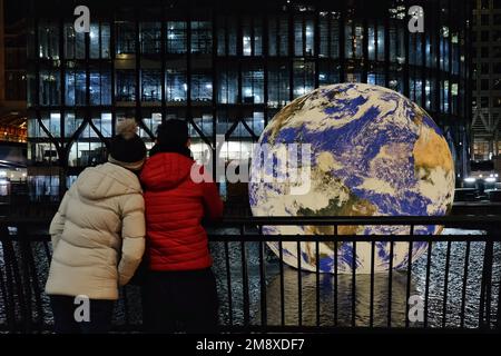 London, Großbritannien. 15. Januar 2023. Ein Paar denkt über die Kunstwerke nach. Der Künstler Luke Jerram's Floating Earth wird vor dem Canary Wharf's Winter Lights Festival installiert, das am 18. Januar eröffnet wird. Die intern beleuchtete Nachbildung der Erde mit einem Durchmesser von zehn Metern wurde aus der Sicht der NASA erstellt und lädt den Zuschauer ein, die Zerbrechlichkeit des Planeten zu erkennen und Umweltbelange zu berücksichtigen. Kredit: Elfte Stunde Fotografie/Alamy Live News Stockfoto