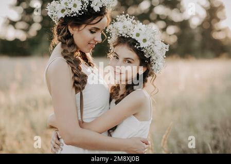 Fröhliche mutter und Tochter in weißen Kleidern mit Blumenkränzen und Bo-Zöpfen im Sommer auf einem Feld in der Natur. Ein kleines Körnchen hinzugefügt, eine Imitation von Fi Stockfoto