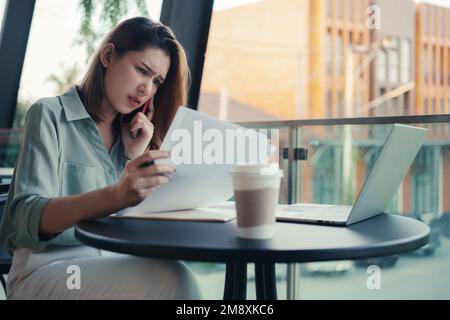 Unzufriedene junge Frau, die Dokumente in der Hand hält und am Handy über ein Problem oder einen Vertrag im Café spricht. Stockfoto