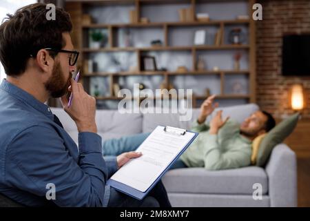 Ein Besuch beim Psychologen eines afroamerikanischen Mannes. Ein gutaussehender Mann liegt auf der Couch und spricht mit einem Psychotherapeuten. Depression, Apathie Stockfoto