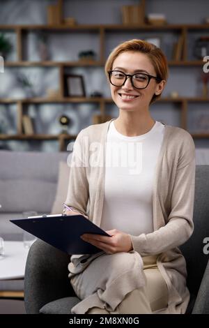 Eine hübsche, lächelnde, selbstbewusste, blonde Frau mit einem schönen Gesicht sieht in die Kamera. Psychologin oder Geschäftsfrau zu Hause im Büro, glücklich Stockfoto