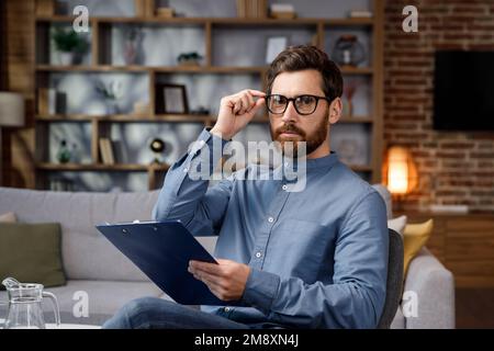 Ein gutaussehender Erwachsener sitzt zu Hause in einem stilvollen Büro. Psychologe, Trainer oder Geschäftsmann, der in die Kamera schaut und lächelt. Schöne Zähne und Stockfoto