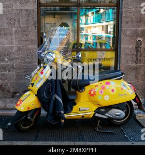 Gelbe Vespa, auch bekannt als Scooter, mit Blumendesign und Lerntellern vor einem Fenster, die ein gelbes Taxi reflektieren. London, England Stockfoto