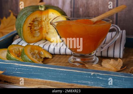 Hausgemachte Kürbismarmelade in einem Glasgefäß auf einem Holztablett mit getrockneten Blättern und geschnittenem Kürbis auf dem Boden Stockfoto