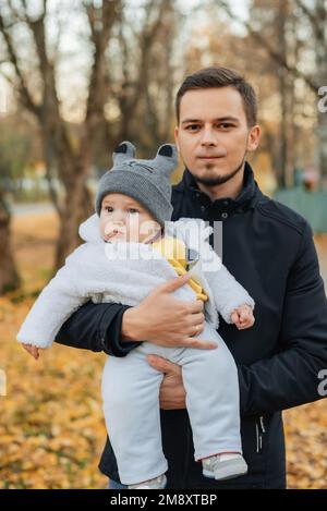 Der junge weiße männliche Vater hält seinen kleinen Sohn im Herbst im Park in den Händen Stockfoto