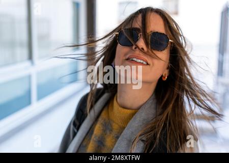 Fröhliche junge Frau in Sonnenbrille und Mantel mit fliegenden dunklen Haaren, die sich auf einem unscharfen Straßenhintergrund drehen Stockfoto