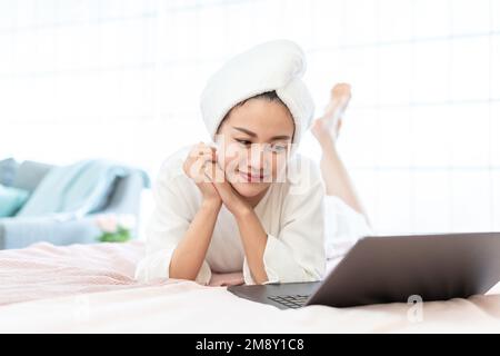 Eine junge Frau, die einen Bademantel trägt, um einen Computer auf dem Bett zu benutzen Stockfoto