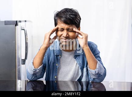 Ein indischer Mann mit einer Brille, die Kopfweh hat, sitzt neben einem Tisch auf weißem Hintergrund Stockfoto