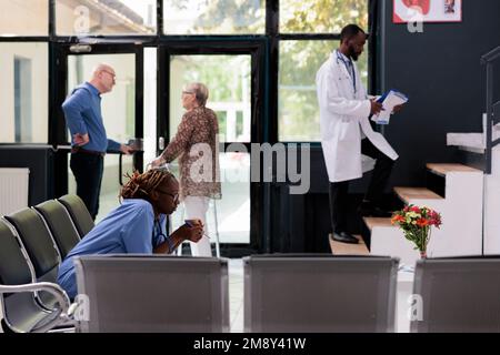 afroamerikanische Krankenschwester, die auf einem Stuhl in der Krankenhauslobby sitzt, während der Konsultation müde ist, eine Pause hat, bevor sie bei der Patientenversorgung zu arbeiten beginnen. Medizinischer Dienst und Konzept Stockfoto