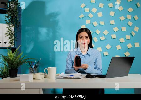 Weibliche Mitarbeiterin an einem modernen Büroschreibtisch mit dunklem Behälter mit Pillen, die auf die Kamera schauen. Professionelle Frau, die eine Kapselflasche mit blauer Wand im Hintergrund zeigt. Stockfoto